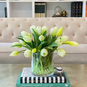 May include: A bouquet of white tulips with green stems in a clear glass vase on a table with a stack of books. The books have black and white stripes and the top book has the text "YVES SAINT LAURENT THE SCANDAL COLLECTION 1971" printed on the cover.