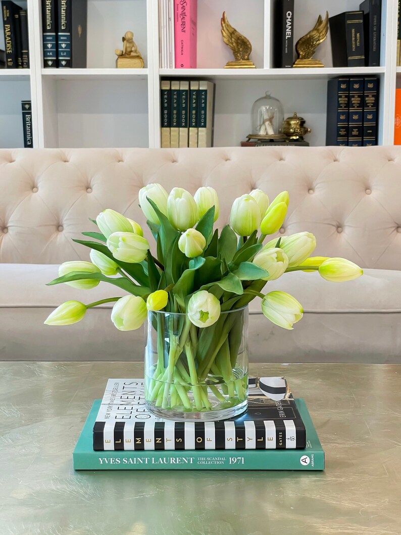 May include: A bouquet of white tulips with green stems in a clear glass vase on a table with a stack of books. The books have black and white stripes and the top book has the text "YVES SAINT LAURENT THE SCANDAL COLLECTION 1971" printed on the cover.