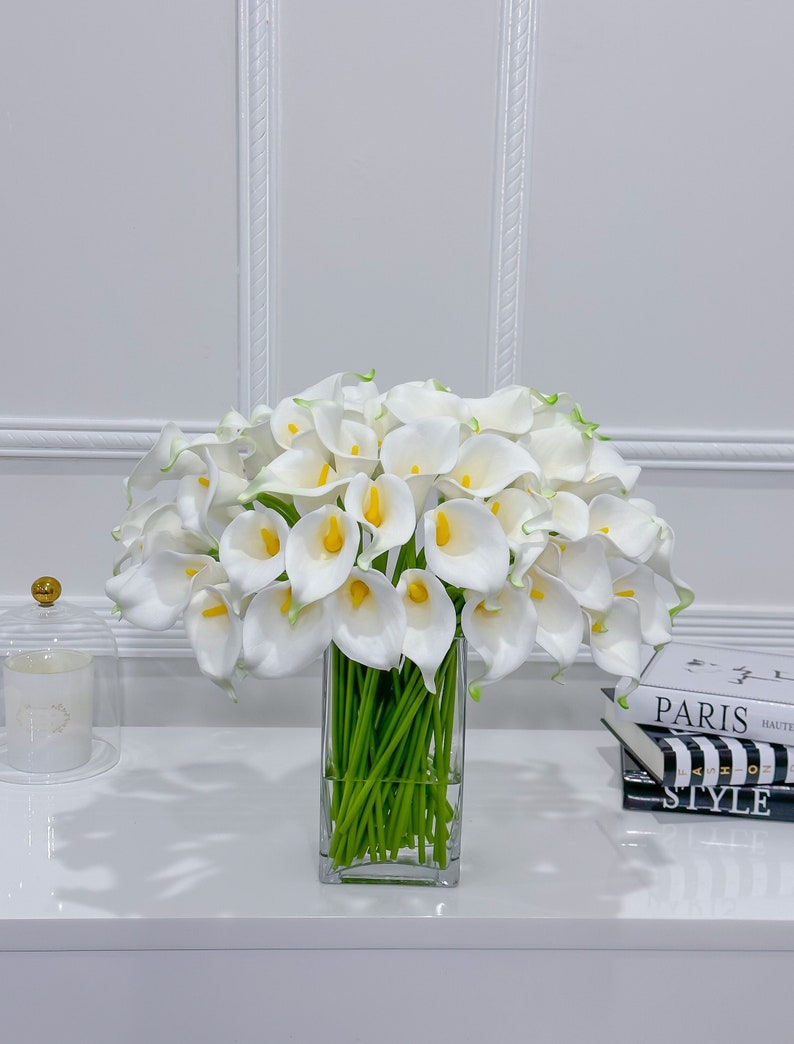 May include: A display of white calla lilies with yellow centres and green stems, presented in a clear, rectangular glass vase. The flowers are arranged on a white surface, with a glass cloche and books in the background.