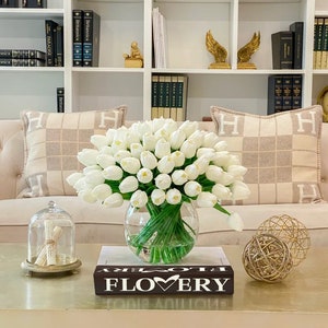 May include: A bouquet of white tulips in a clear glass vase on a coffee table with a book that says "FLOVERY" on the cover. The table is in front of a white couch with two pillows with an "H" monogram.