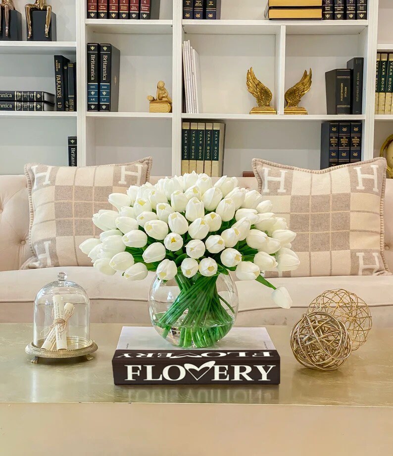 May include: A bouquet of white tulips in a clear glass vase on a coffee table with a book that says "FLOVERY" on the cover. The table is in front of a white couch with two pillows with an "H" monogram.