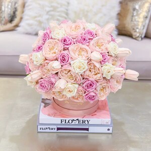 Puede incluir: Un arreglo floral rosa y blanco en una caja rosa, sentado encima de dos libros con la palabra "FLOWERY" impresa en la portada.