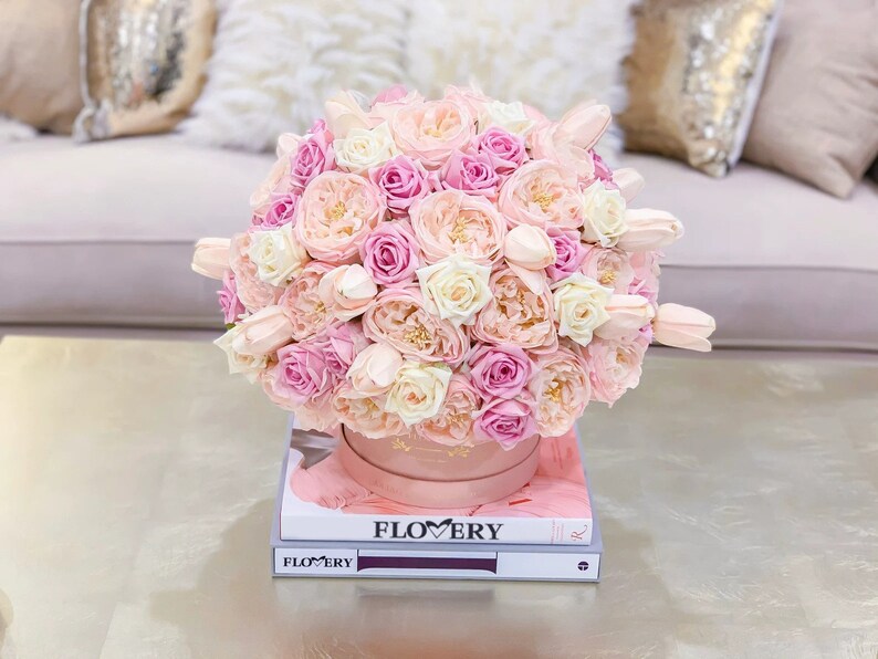 Puede incluir: Un arreglo floral rosa y blanco en una caja rosa, sentado encima de dos libros con la palabra "FLOWERY" impresa en la portada.