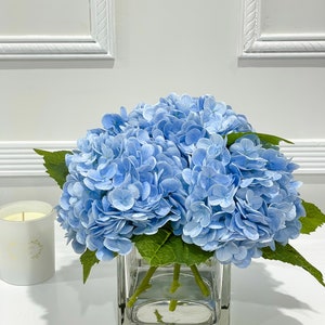 May include: A bouquet of light blue hydrangeas in a clear glass vase. The flowers are arranged in a simple, elegant style, and the vase is square shaped.