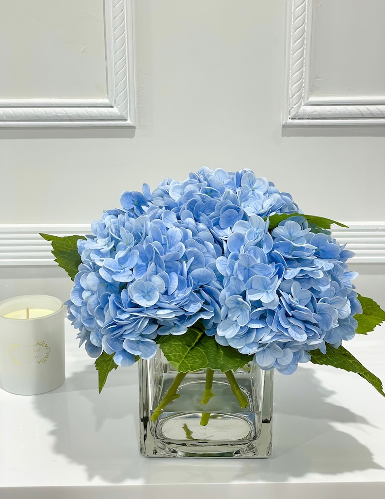 May include: A bouquet of light blue hydrangeas in a clear glass vase. The flowers are arranged in a simple, elegant style, and the vase is square shaped.
