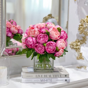 K&ouml;nnte beinhalten: Ein Strau&szlig; rosa Pfingstrosen in einer klaren Glasvase auf zwei B&uuml;chern. Das obere Buch tr&auml;gt den Titel "Live Beautiful" und das untere Buch den Titel "White".