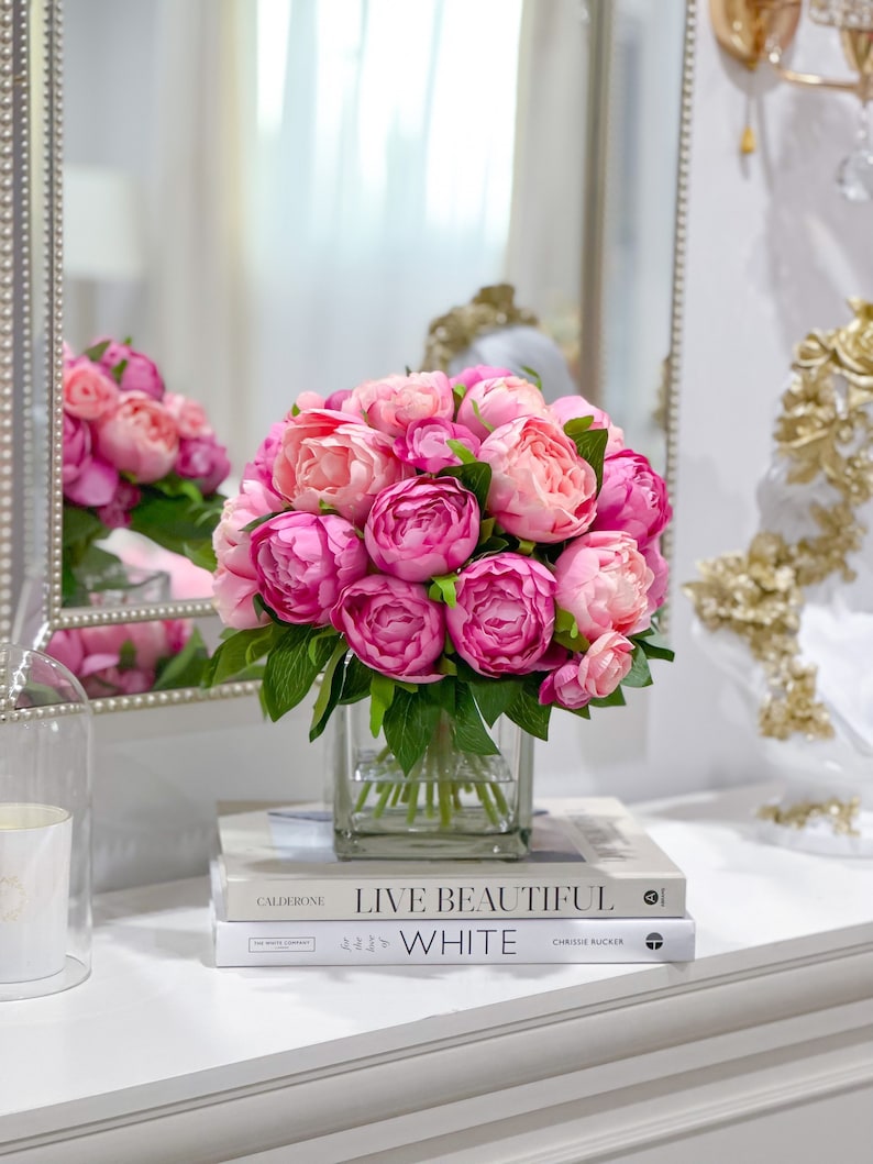 K&ouml;nnte beinhalten: Ein Strau&szlig; rosa Pfingstrosen in einer klaren Glasvase auf zwei B&uuml;chern. Das obere Buch tr&auml;gt den Titel "Live Beautiful" und das untere Buch den Titel "White".