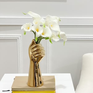 May include: A gold-colored ceramic vase shaped like a raised fist holding a bouquet of white calla lilies. The vase is on a white table with a stack of three books.