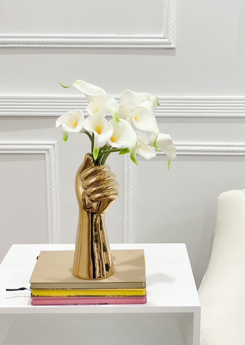 May include: A gold-colored ceramic vase shaped like a raised fist holding a bouquet of white calla lilies. The vase is on a white table with a stack of three books.