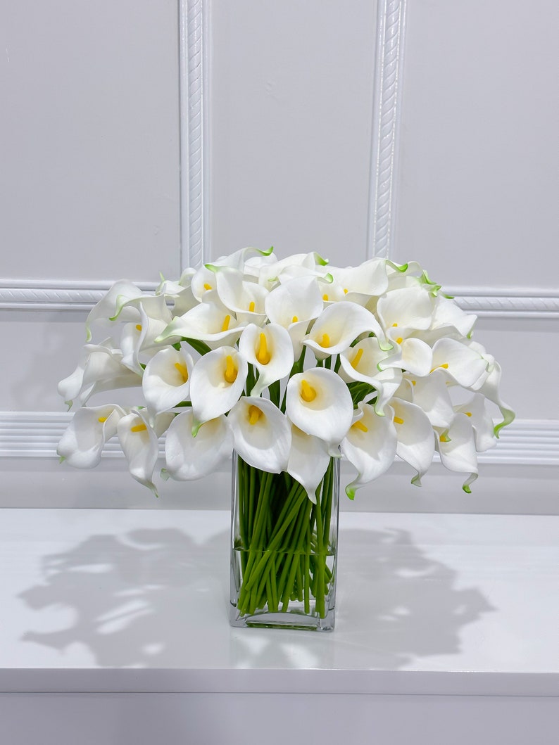 May include: A display of white calla lilies with yellow centres and green tips, presented in a clear, square glass vase. The flowers are set against a white backdrop, creating a clean and elegant aesthetic. The vase rests on a white surface.