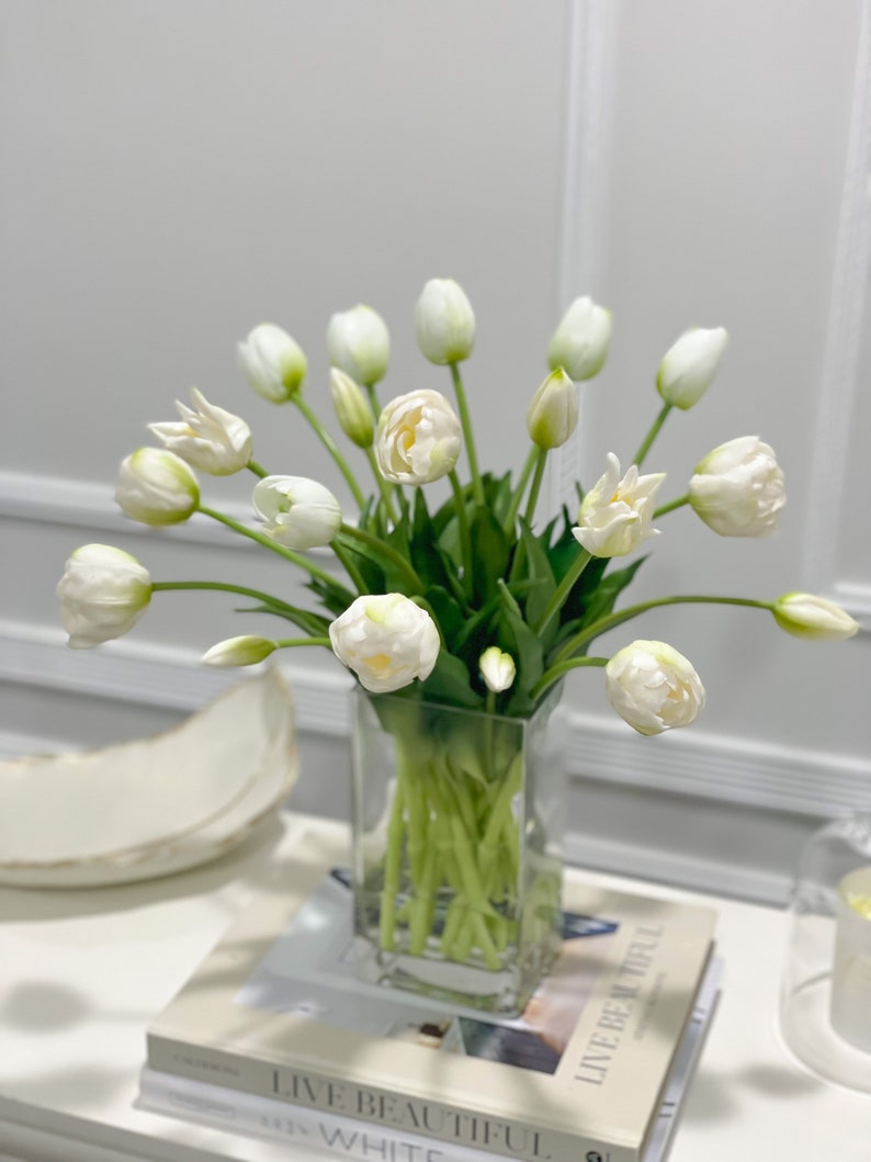 Puede incluir: Un ramo de tulipanes blancos en un jarr&oacute;n de vidrio transparente. El jarr&oacute;n est&aacute; sobre una pila de libros con el texto "Live Beautiful" en el libro de arriba.