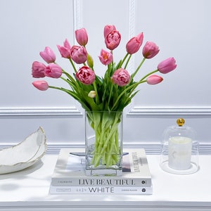 Puede incluir: Un ramo de tulipanes rosas en un jarr&oacute;n de vidrio transparente sobre una mesa blanca con dos libros, uno titulado "Live Beautiful White" de Chrissie Rucker y el otro titulado "Calderone" de The New York Times.