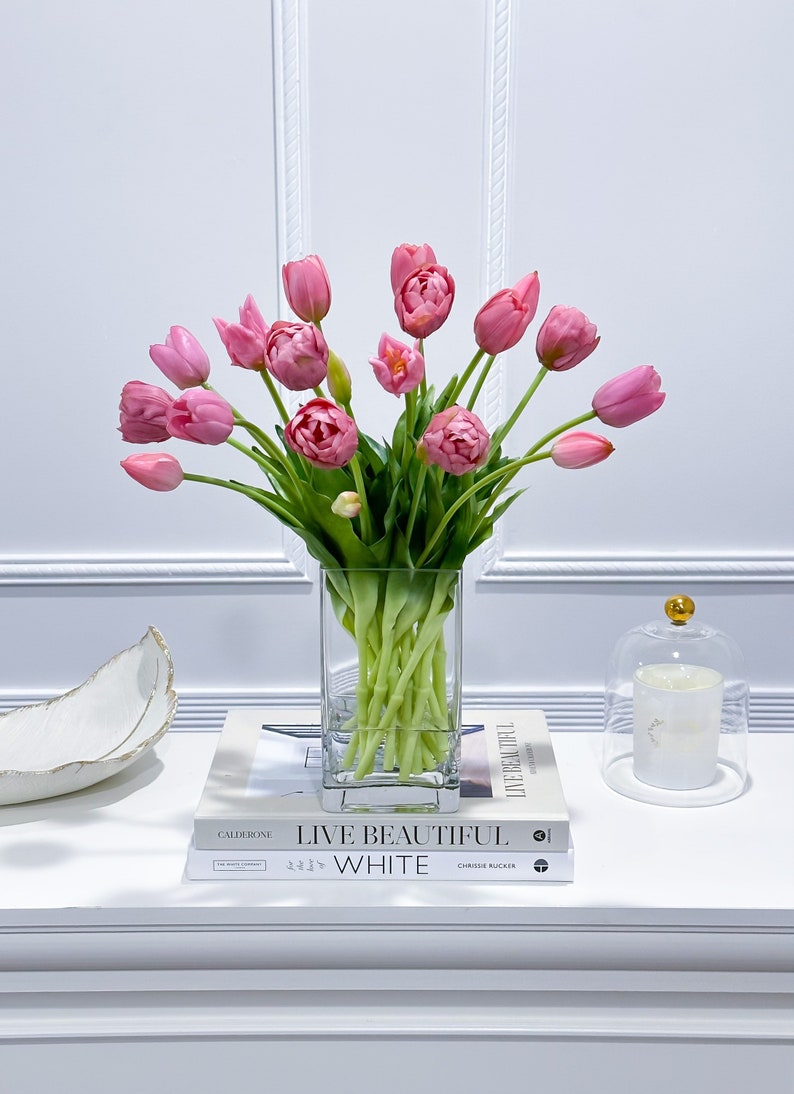 Puede incluir: Un ramo de tulipanes rosas en un jarr&oacute;n de vidrio transparente sobre una mesa blanca con dos libros, uno titulado "Live Beautiful White" de Chrissie Rucker y el otro titulado "Calderone" de The New York Times.