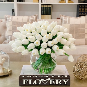 May include: A bouquet of white artificial tulips in a clear glass vase on a coffee table with a book that says "FLOWERY" on the cover. The vase is sitting on a book with the word "FLOWERY" on the cover. The tulips are arranged in a vase on a coffee table.