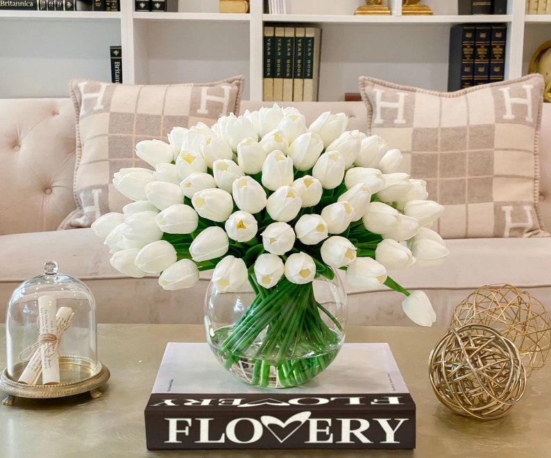 May include: A bouquet of white artificial tulips in a clear glass vase on a coffee table with a book that says "FLOWERY" on the cover. The vase is sitting on a book with the word "FLOWERY" on the cover. The tulips are arranged in a vase on a coffee table.
