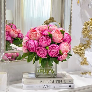 K&ouml;nnte beinhalten: Ein Strau&szlig; rosa Pfingstrosen in einer klaren Glasvase, die auf zwei B&uuml;chern steht. Das obere Buch tr&auml;gt den Titel "Live Beautiful" und das untere Buch den Titel "White".