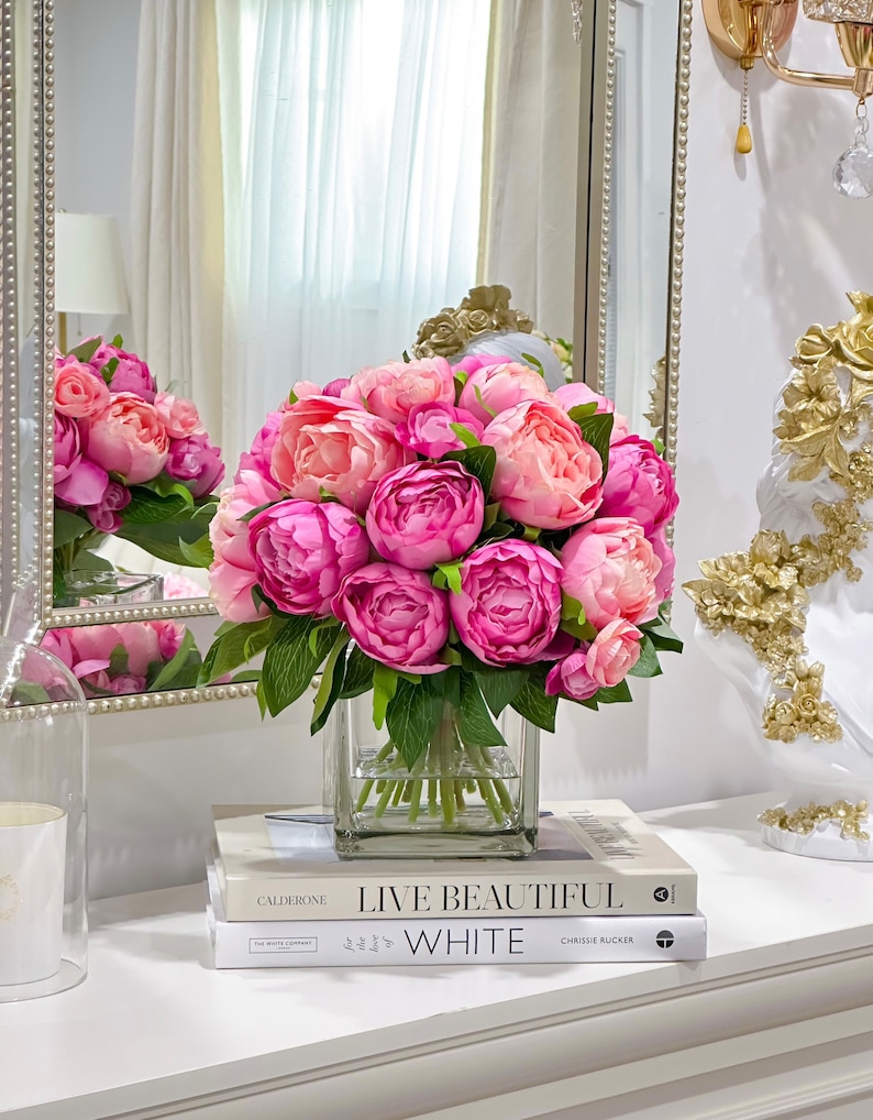 K&ouml;nnte beinhalten: Ein Strau&szlig; rosa Pfingstrosen in einer klaren Glasvase, die auf zwei B&uuml;chern steht. Das obere Buch tr&auml;gt den Titel "Live Beautiful" und das untere Buch den Titel "White".