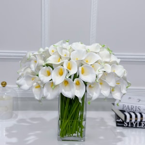May include: A vibrant display of white calla lilies with yellow centres and green stems, presented in a clear, rectangular glass vase. The flowers are set against a white backdrop, creating a clean and elegant aesthetic. Decorative books are visible in the background.