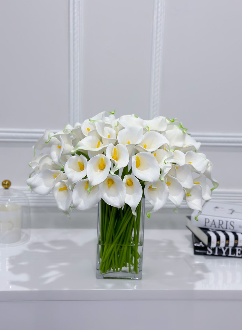 May include: A vibrant display of white calla lilies with yellow centres and green stems, presented in a clear, rectangular glass vase. The flowers are set against a white backdrop, creating a clean and elegant aesthetic. Decorative books are visible in the background.