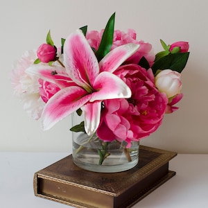 May include: A bouquet of pink and white peonies and a pink lily in a clear glass vase. The vase is sitting on a brown book with gold accents.