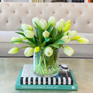 May include: A bouquet of white and green tulips in a clear glass vase on a table with books stacked underneath. The books have black and white stripes and the text "Elements of Style" and "Yves Saint Laurent The Scandal Collection 1971".