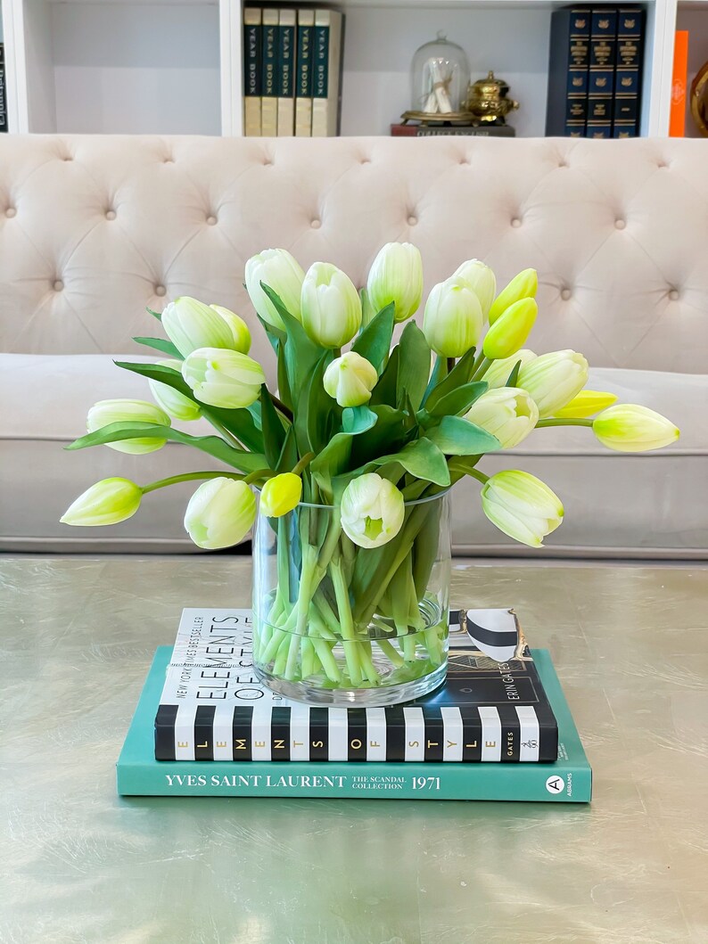 May include: A bouquet of white and green tulips in a clear glass vase on a table with books stacked underneath. The books have black and white stripes and the text "Elements of Style" and "Yves Saint Laurent The Scandal Collection 1971".