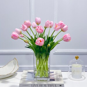 Puede incluir: Un ramo de tulipanes rosas en un jarr&oacute;n de vidrio transparente sobre una mesa blanca con dos libros. Los libros se titulan "Live Beautiful" y "White".