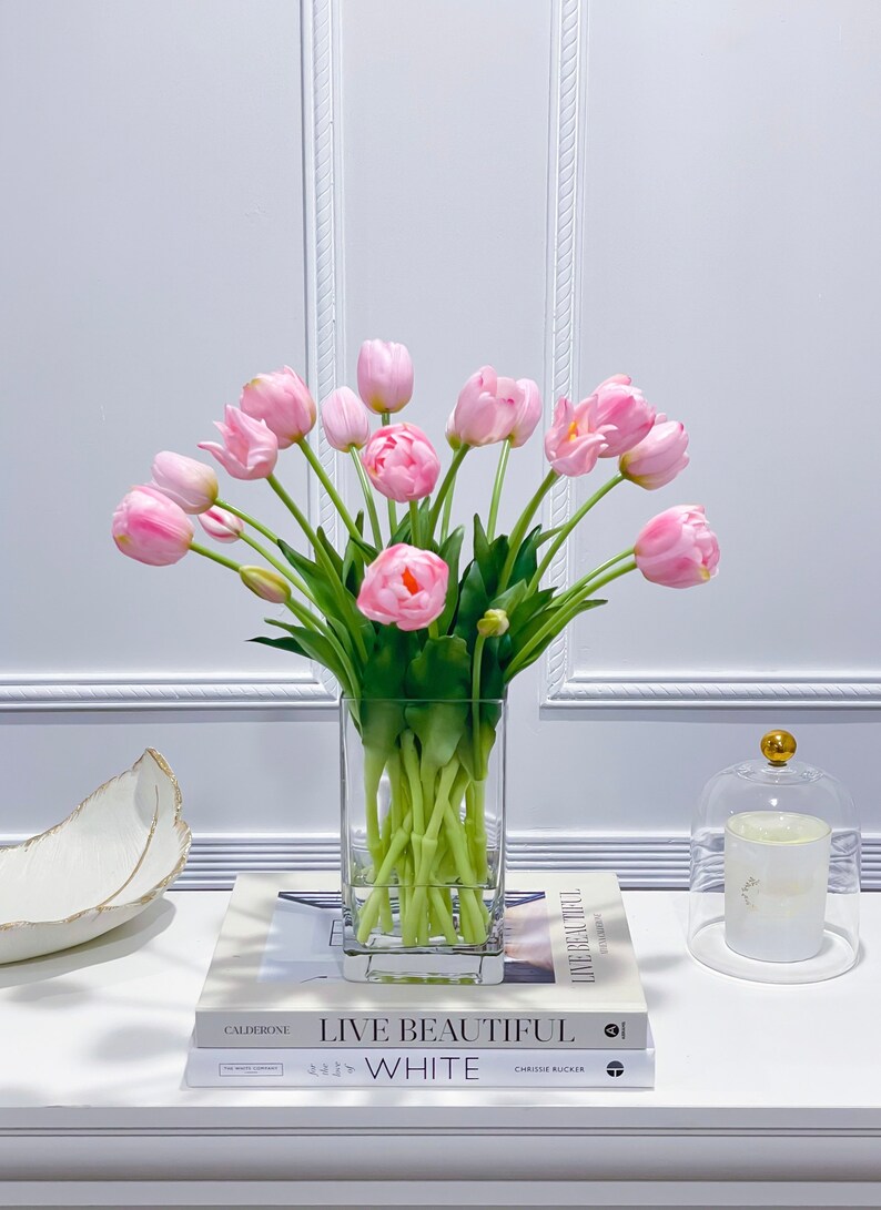 Puede incluir: Un ramo de tulipanes rosas en un jarr&oacute;n de vidrio transparente sobre una mesa blanca con dos libros. Los libros se titulan "Live Beautiful" y "White".