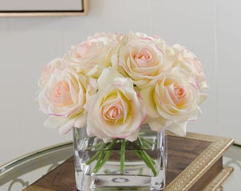 May include: A bouquet of cream-colored roses with pink edges, arranged in a clear glass cube vase. The roses are in a cluster, with the stems visible in the vase.