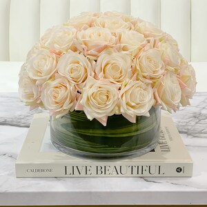 May include: A bouquet of blush pink roses arranged in a clear glass vase with green stripes. The vase is sitting on a white marble table with a book titled "Live Beautiful" by Calderone.