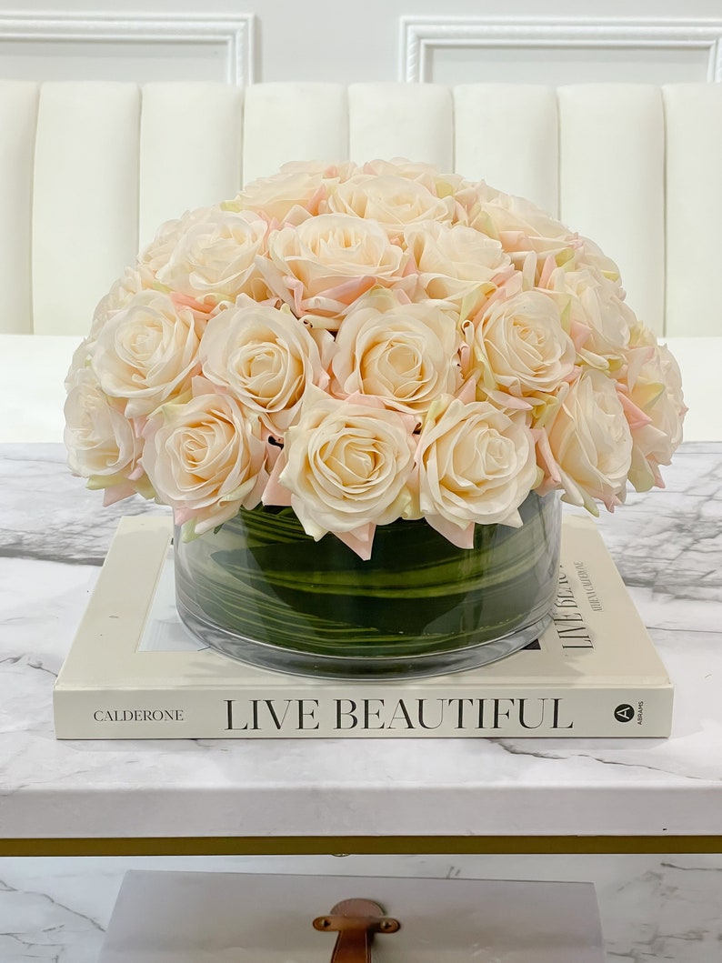May include: A bouquet of blush pink roses arranged in a clear glass vase with green stripes. The vase is sitting on a white marble table with a book titled "Live Beautiful" by Calderone.