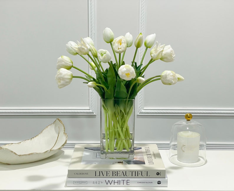 Puede incluir: Un jarr&oacute;n de vidrio transparente lleno de tulipanes blancos. El jarr&oacute;n est&aacute; sobre una mesa blanca con dos libros y una vela blanca en una campana de vidrio.