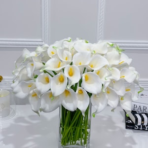 May include: A large bouquet of white calla lilies with yellow centres and green stems in a clear, square glass vase. The flowers have a slight green edge. The arrangement sits on a white surface.