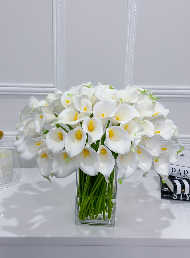 May include: A large bouquet of white calla lilies with yellow centres and green stems in a clear, square glass vase. The flowers have a slight green edge. The arrangement sits on a white surface.