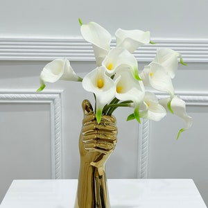 May include: A gold-colored ceramic hand vase with a bouquet of white calla lilies.