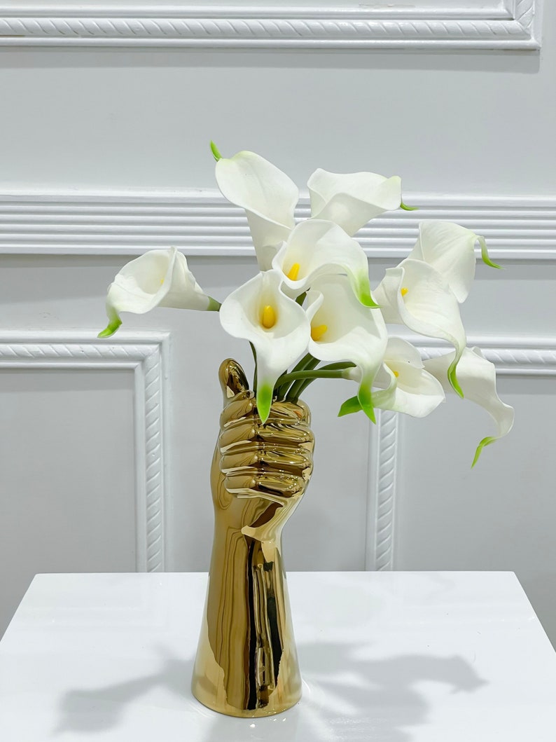 May include: A gold-colored ceramic hand vase with a bouquet of white calla lilies.