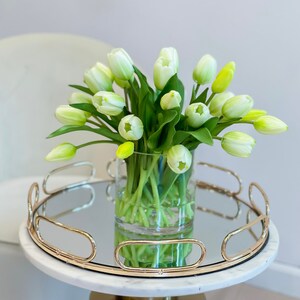 May include: A bouquet of white tulips in a clear glass vase on a gold mirrored tray with a white marble base.