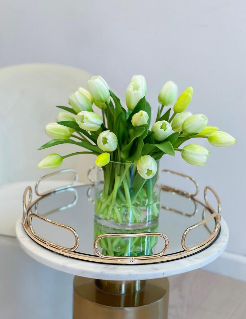 May include: A bouquet of white tulips in a clear glass vase on a gold mirrored tray with a white marble base.