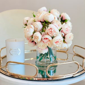May include: A white candle with gold lettering that reads "The Roses Flowery" sits on a gold mirrored tray with a bouquet of pale pink roses in a clear glass vase.