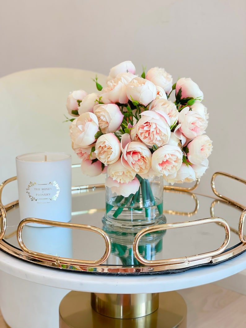 May include: A white candle with gold lettering that reads "The Roses Flowery" sits on a gold mirrored tray with a bouquet of pale pink roses in a clear glass vase.