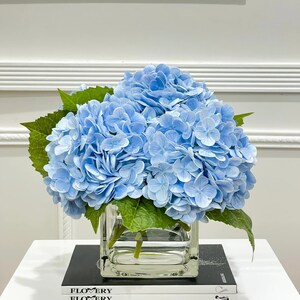 May include: A bouquet of light blue hydrangeas in a clear glass cube vase, sitting on a stack of books with the word "FLOWERY" printed on the covers.