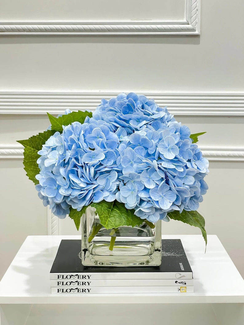 May include: A bouquet of light blue hydrangeas in a clear glass cube vase, sitting on a stack of books with the word "FLOWERY" printed on the covers.