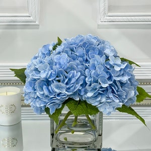 May include: A bouquet of light blue hydrangeas in a clear glass cube vase. The vase is sitting on a white surface.