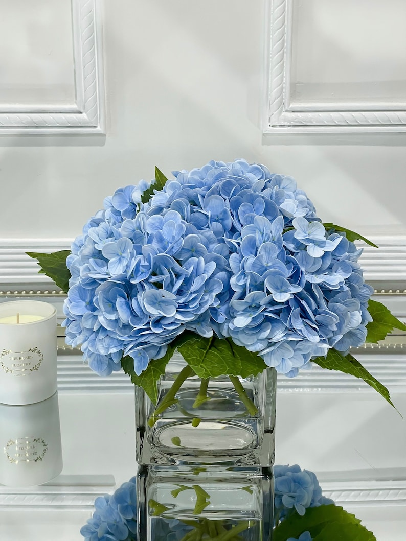 May include: A bouquet of light blue hydrangeas in a clear glass cube vase. The vase is sitting on a white surface.