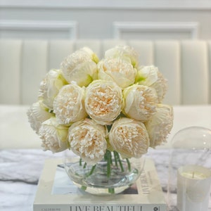 May include: A bouquet of white peonies in a clear glass vase, sitting on top of two books. The top book is titled "Live Beautiful White" by  Chassidic Ruckser. The bottom book is titled "Calderone".