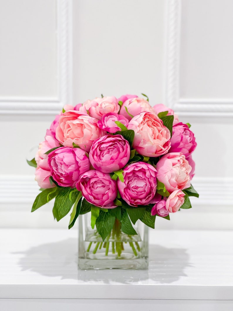 K&ouml;nnte beinhalten: Ein Strau&szlig; rosa Pfingstrosen, die in einer klaren Glasvase arrangiert sind. Die Blumen sind in voller Bl&uuml;te und haben ein weiches, zartes Aussehen.
