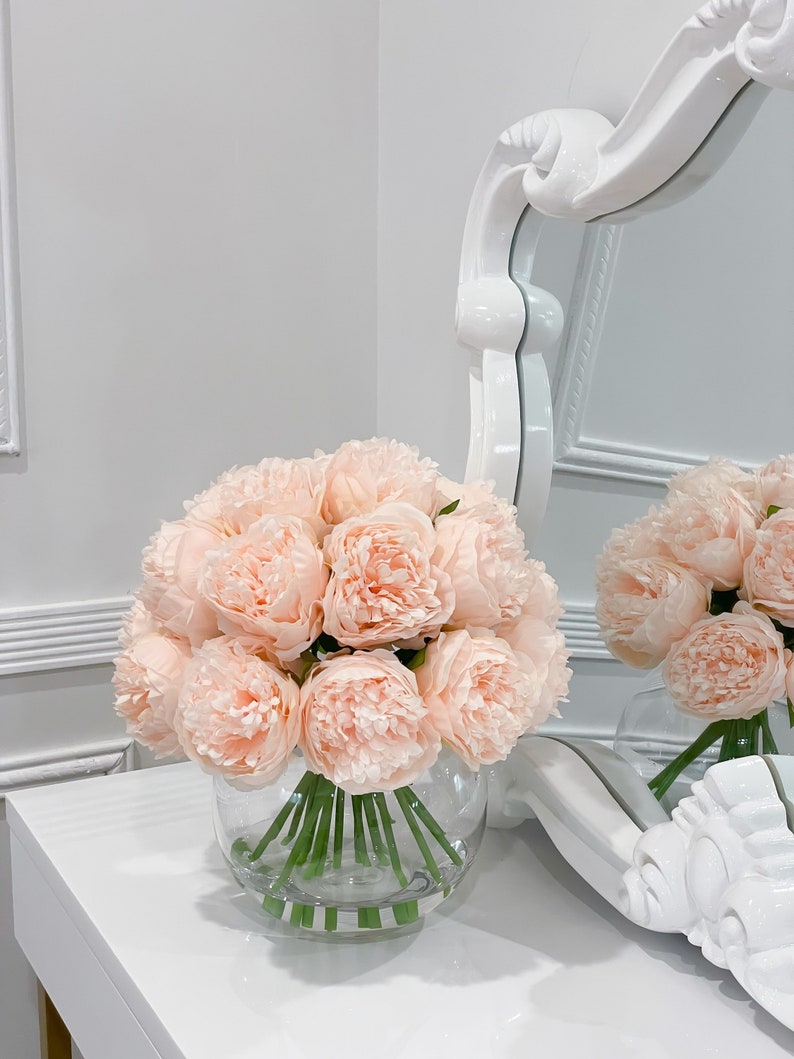 May include: A bouquet of pale pink peonies arranged in a clear glass vase. The flowers are on a white surface with a white ornate mirror in the background.