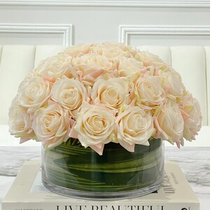 May include: A bouquet of cream-colored roses with pink edges arranged in a clear glass vase with green stripes. The vase is sitting on a white book with the text "Live Beautiful" printed on the cover.