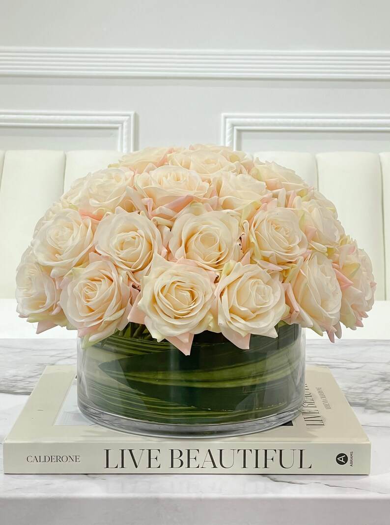 May include: A bouquet of cream-colored roses with pink edges arranged in a clear glass vase with green stripes. The vase is sitting on a white book with the text "Live Beautiful" printed on the cover.