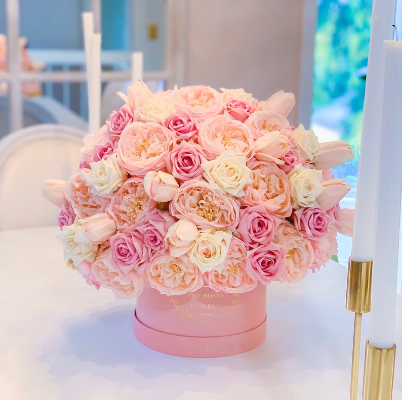 Puede incluir: Una caja rosa llena de un gran ramo de rosas rosas y blancas. La caja tiene un logotipo dorado que dice "The Roses Flowery".